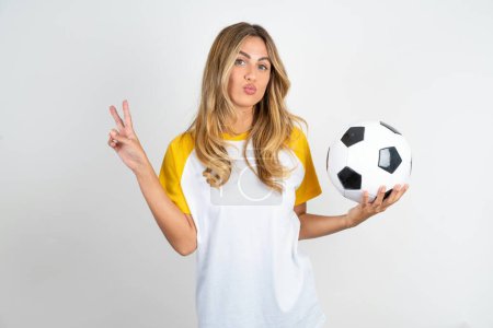 belle femme portant le football T-shirt sur fond blanc fait geste de paix garde les lèvres pliées montre v signe tenant ballon de football. Concept de langage corporel