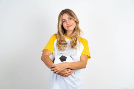 Foto de Mujer hermosa y segura con camiseta de fútbol sobre fondo blanco con pelota de fútbol mirando a la cámara - Imagen libre de derechos