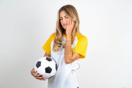 Foto de Hermosa mujer con camiseta de fútbol sobre fondo blanco con dolor de muelas y pelota de fútbol - Imagen libre de derechos