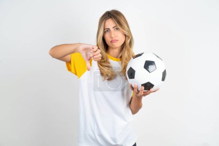 Foto de Hermosa mujer vistiendo camiseta de fútbol sobre fondo blanco mostrando el pulgar hacia abajo signo sosteniendo pelota de fútbol - Imagen libre de derechos