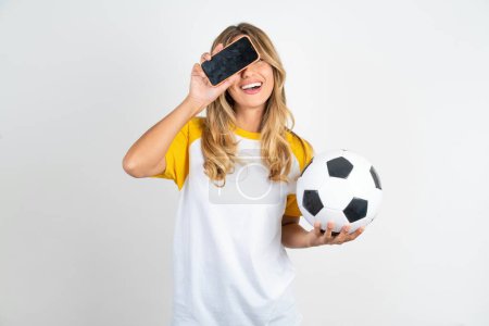 Photo for Beautiful woman wearing football T-shirt over white background holding modern smartphone covering one eye while smiling and holding  soccer ball - Royalty Free Image