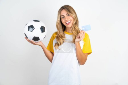 Téléchargez les photos : Portrait de belle femme portant un T-shirt de football sur fond blanc tenant carte de crédit et ballon de football - en image libre de droit