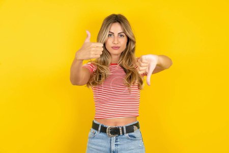 Photo for Beautiful blonde young woman wearing striped t-shirt over yellow studio background showing thumb up and thumb down, difficult choose concept - Royalty Free Image