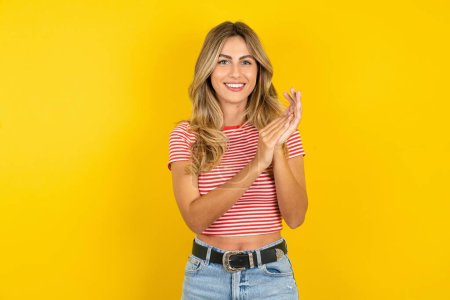Téléchargez les photos : Joyeux et joyeux jeune femme portant t-shirt rayé sur fond jaune studio applaudissements mains - en image libre de droit