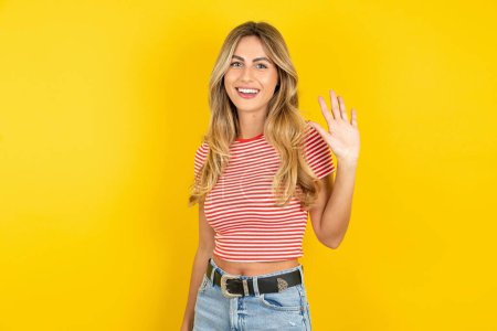Téléchargez les photos : Belle jeune femme blonde portant un t-shirt rayé sur fond jaune studio agite sa main en saluant et en souriant, geste de bienvenue amical. - en image libre de droit