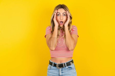 Photo for Beautiful blonde young woman wearing striped t-shirt over yellow studio background with scared expression, keeps hands on head, jaw dropped, has terrific expression. Omg concept - Royalty Free Image