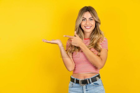 Foto de Hermosa mujer joven rubia con camiseta a rayas sobre fondo amarillo estudio señalando y sosteniendo la mano mostrando anuncios - Imagen libre de derechos