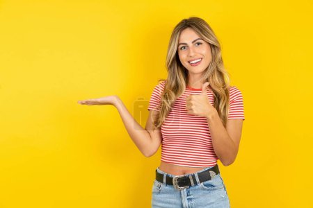 Photo for Beautiful blonde young woman wearing striped t-shirt over yellow studio background showing palm hand and doing ok gesture with thumbs up, smiling happy and cheerful. - Royalty Free Image