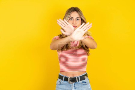 Téléchargez les photos : Jeune femme blonde portant un t-shirt rayé sur fond jaune studio a l'expression de rejet traversant les bras et les paumes faisant signe négatif, visage en colère. - en image libre de droit