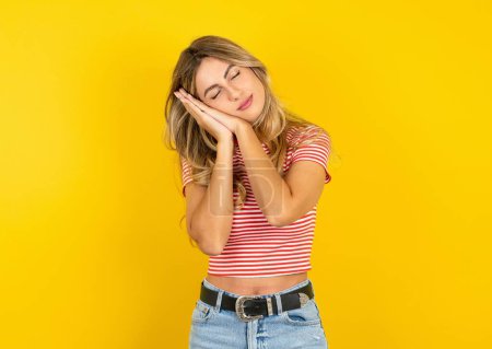 Téléchargez les photos : Belle jeune femme blonde portant un t-shirt rayé sur fond jaune studio dormant fatigué rêvant et posant les mains ensemble tout en souriant les yeux fermés. - en image libre de droit