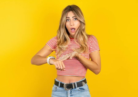 Téléchargez les photos : Belle jeune femme blonde portant un t-shirt rayé sur fond jaune studio à la hâte pointant vers regarder le temps, l'impatience, contrarié et en colère pour retard de délai. - en image libre de droit