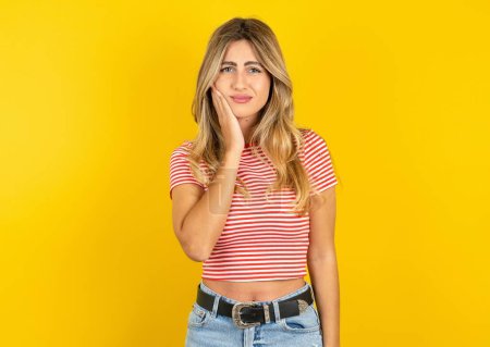 Photo for Young beautiful blonde woman wearing striped t-shirt over yellow studio background touching mouth with hand with painful expression because of toothache or dental illness on teeth. - Royalty Free Image