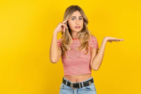 Photo for Beautiful blonde young woman wearing striped t-shirt over yellow studio background confused and annoyed with open palm showing copy space and pointing finger to forehead. Think about it. - Royalty Free Image