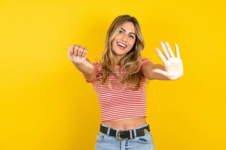 Foto de Hermosa mujer rubia con camiseta a rayas sobre fondo amarillo estudio mostrando y apuntando hacia arriba con los dedos número seis mientras sonríe confiado y feliz. - Imagen libre de derechos