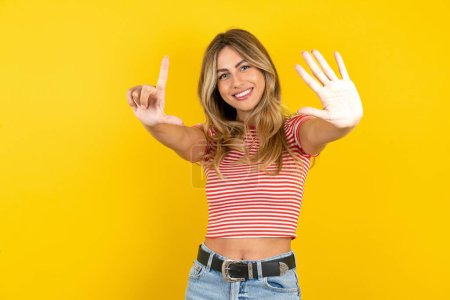 Foto de Hermosa mujer joven rubia con camiseta a rayas sobre fondo amarillo estudio mostrando y apuntando hacia arriba con los dedos número siete mientras sonríe confiado y feliz. - Imagen libre de derechos