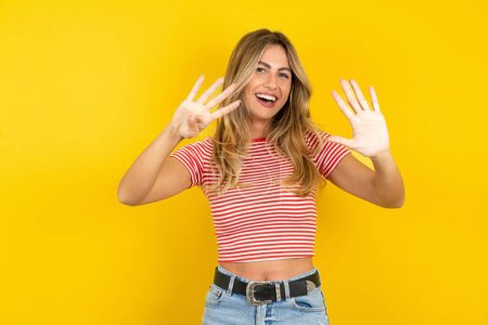 Téléchargez les photos : Jeune belle femme blonde portant un t-shirt rayé sur fond jaune studio montrant et pointant vers le haut avec les doigts numéro neuf tout en souriant confiant et heureux. - en image libre de droit