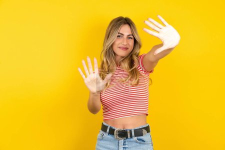 Téléchargez les photos : Jeune belle femme blonde portant un t-shirt rayé sur fond jaune studio montrant et pointant vers le haut avec les doigts numéro dix tout en souriant confiant et heureux. - en image libre de droit