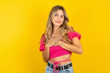 Photo for Beautiful blonde young woman wearing pink crop top on yellow background smiling with hands on chest and grateful gesture on face. Health concept. - Royalty Free Image