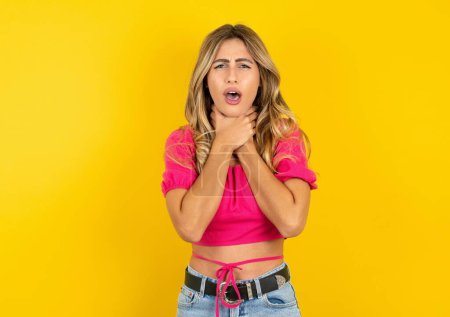 Photo for Young blonde woman wearing pink crop top on yellow background shouting suffocate because painful strangle. Health problem. Asphyxiate and suicide concept. - Royalty Free Image