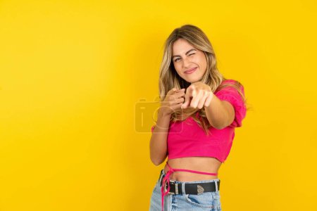 Foto de Joven rubia alegre vistiendo top rosa sobre guiños de fondo amarillo y señala los dedos índice a la cámara, elige a alguien, hace pistola de dedo. - Imagen libre de derechos