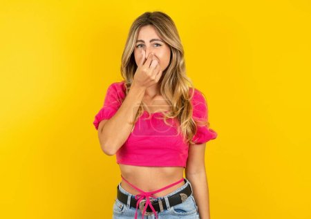 Photo for Beautiful blonde young woman wearing pink crop top on yellow background smelling something stinky and disgusting, intolerable smell, holding breath with fingers on nose. Bad smell - Royalty Free Image