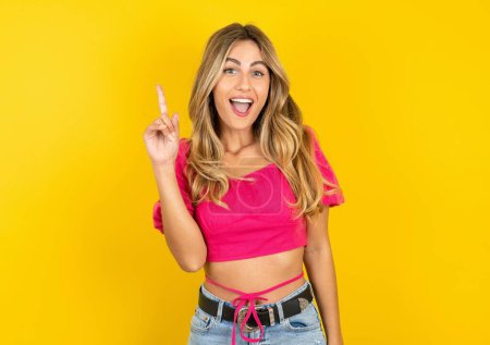 Photo for Beautiful blonde young woman wearing pink crop top on yellow background pointing finger up and looking inspired by genius thought, showing good idea sign, having clever solution in mind - Royalty Free Image