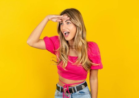 Photo for Very happy and smiling blonde young woman wearing pink crop top on yellow background looking far away with hand over head. Searching concept. - Royalty Free Image