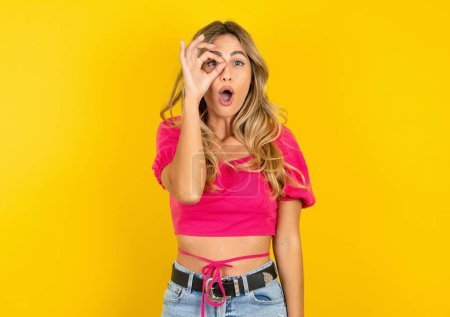 Photo for Beautiful blonde young woman wearing pink crop top on yellow background doing ok gesture shocked with surprised face, eye looking through fingers. Unbelieving expression - Royalty Free Image