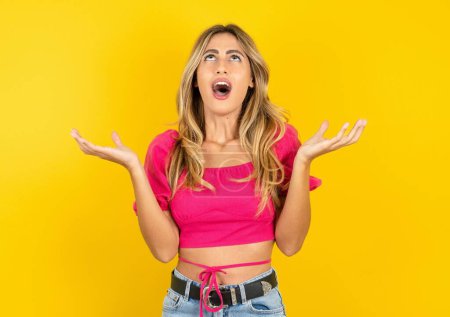 Photo for Young blonde woman wearing pink crop top on yellow background crazy and mad shouting and yelling with aggressive expression and arms raised. Frustration concept. - Royalty Free Image