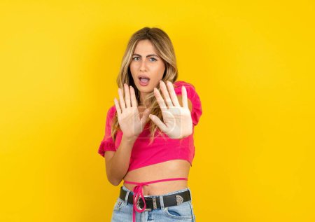Photo for Young blonde woman wearing pink crop top on yellow background afraid and terrified with fear expression stop gesture with hands, shouting in shock. Panic concept. - Royalty Free Image