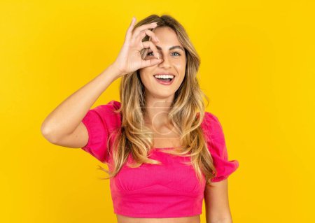 Photo for Blonde young woman wearing pink crop top on yellow background doing ok gesture with hand smiling, eye looking through fingers with happy face. - Royalty Free Image