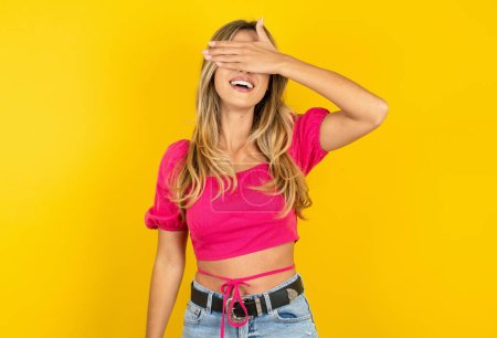 Photo for Blonde young woman wearing pink crop top on yellow background smiling and laughing with hand on face covering eyes for surprise. Blind concept. - Royalty Free Image