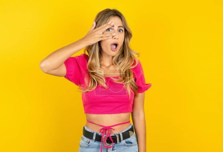 Photo for Young blonde woman wearing pink crop top on yellow background peeking in shock covering face with hand, looking through fingers with embarrassed expression. - Royalty Free Image