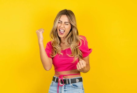 Téléchargez les photos : Très heureuse et excitée jeune femme blonde portant un haut de culture rose sur fond jaune faisant geste gagnant avec les bras levés, souriant et criant pour le succès. Concept de célébration. - en image libre de droit