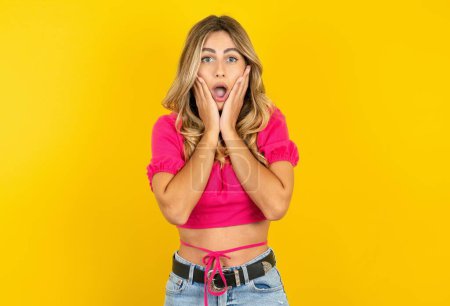Photo for Shocked blonde woman wearing pink crop top on yellow background looks with great surprisement being very stunned, astonished with unexpected news. Facial expressions concept. - Royalty Free Image