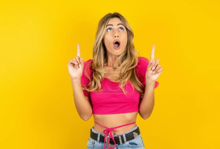 Photo for Amazed and surprised  blonde young woman wearing pink crop top on yellow background looking up and pointing with fingers and raised arms. - Royalty Free Image