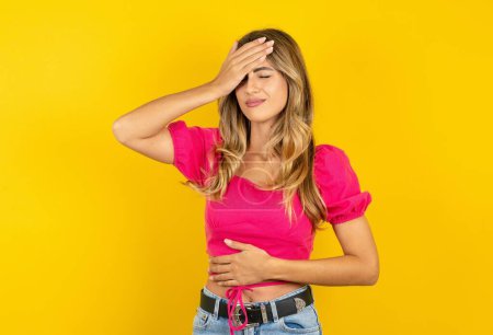Photo for Young blonde woman wearing pink crop top on yellow background touching forehead for illness and fever, flu and cold, virus sick. - Royalty Free Image