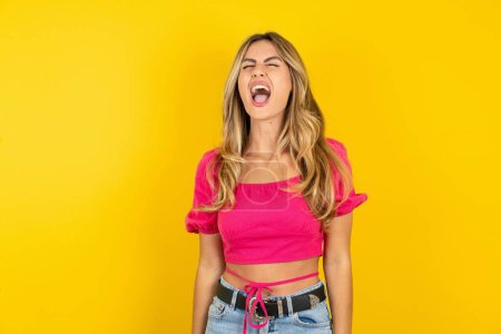 Photo for Angry and mad blonde young woman wearing pink crop top on yellow background screaming frustrated and furious, shouting with anger. Rage and aggressive concept. - Royalty Free Image
