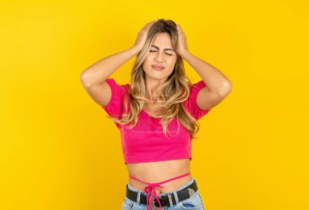 Photo for Young blonde woman wearing pink crop top on yellow background suffering from strong headache desperate and stressed because of overwork. Depression and pain concept. - Royalty Free Image