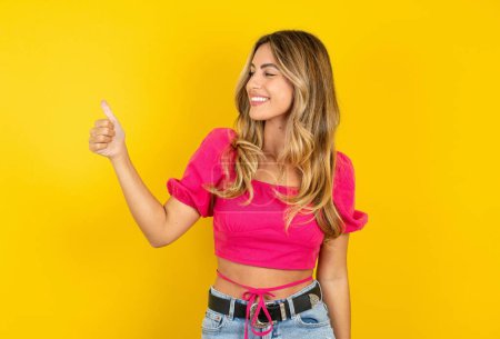 Photo for Beautiful blonde young woman wearing pink crop top on yellow background looking proud, smiling and doing thumbs up gesture to the side. Good job! - Royalty Free Image
