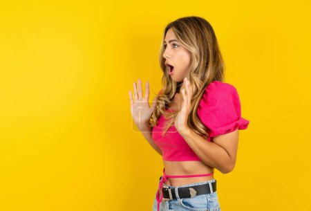 Photo for Blonde young woman wearing pink crop top on yellow background shouts loud, keeps eyes opened and hands tense. - Royalty Free Image