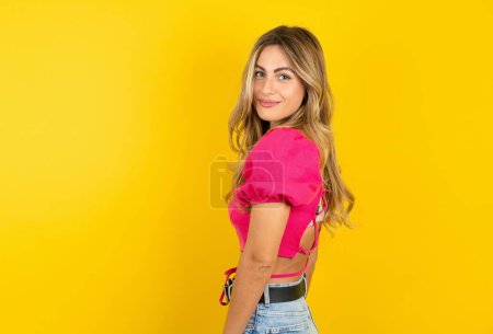Téléchargez les photos : Satisfait jolie jeune femme blonde portant un haut de culture rose sur fond jaune souriant et regardant la caméra - en image libre de droit