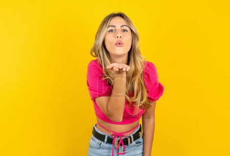 Photo for Young blonde woman wearing pink crop top on yellow background looking at the camera blowing a kiss with hand on air being lovely and sexy. Love expression. - Royalty Free Image