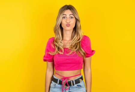 Photo for Shot of pleasant looking blonde young woman wearing pink crop top on yellow background pouts lips, looks at camera. Human facial expressions - Royalty Free Image