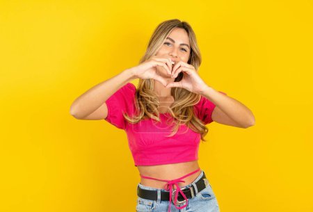 Photo for Beautiful blonde young woman wearing pink crop top on yellow background smiling in love doing heart symbol shape with hands. Romantic concept. - Royalty Free Image