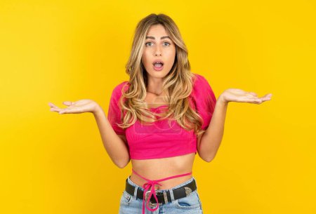 Photo for Frustrated blonde young woman wearing pink crop top on yellow background feels puzzled and hesitant, shrugs shoulders in bewilderment, keeps mouth widely opened, doesn't know what to do. - Royalty Free Image