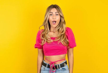 Photo for Oh my God. Surprised blonde young woman wearing pink crop top on yellow background stares at camera with shocked expression exclaims with unexpectedness, - Royalty Free Image
