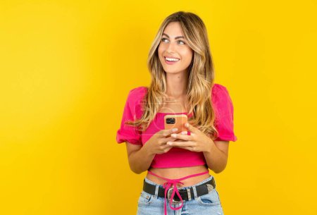 Téléchargez les photos : Belle jeune femme blonde portant un haut de culture rose sur fond jaune tenant un smartphone et regardant de côté l'espace de copie vierge. - en image libre de droit