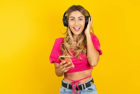 Photo for Positive blonde young woman wearing pink crop top on yellow background holds modern smartphone connected to headphones, clenches fist from good emotions, exclaims with joy - Royalty Free Image