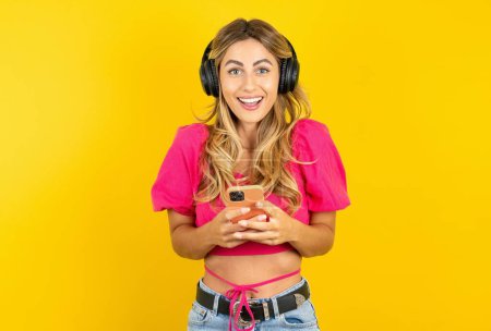 Photo for Excited blonde young woman wearing pink crop top on yellow background holding smartphone and looking amazed to the camera after receiving good news. - Royalty Free Image